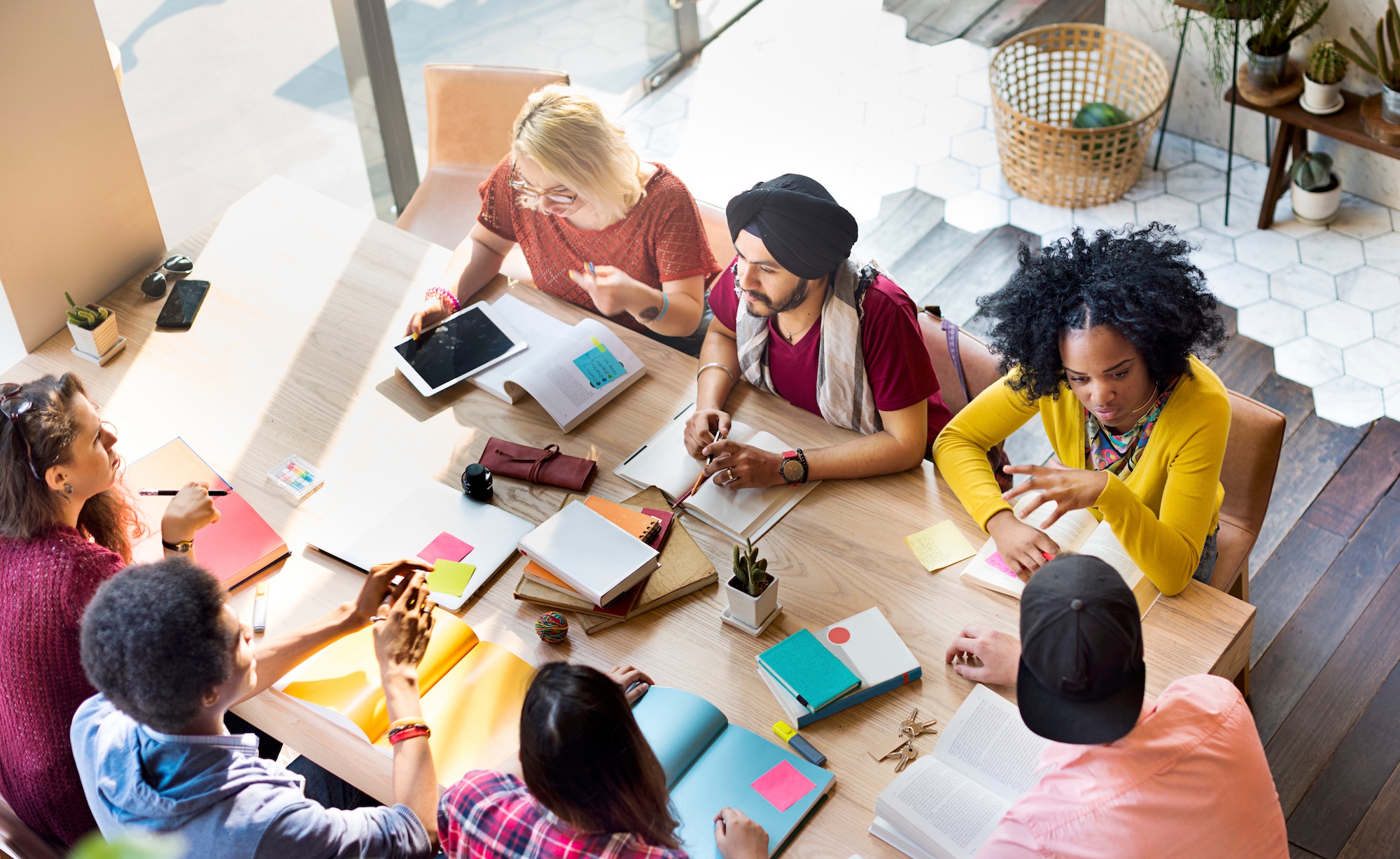 Seven diverse people sitting around a table in a meeting talking with notebooks and a tablet