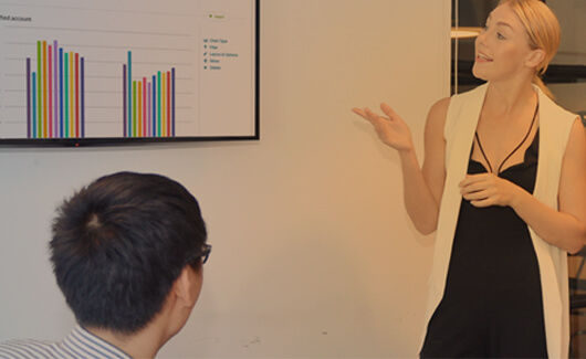 One person standing presenting a bar chart on a screen with another person sitting looking at the screen