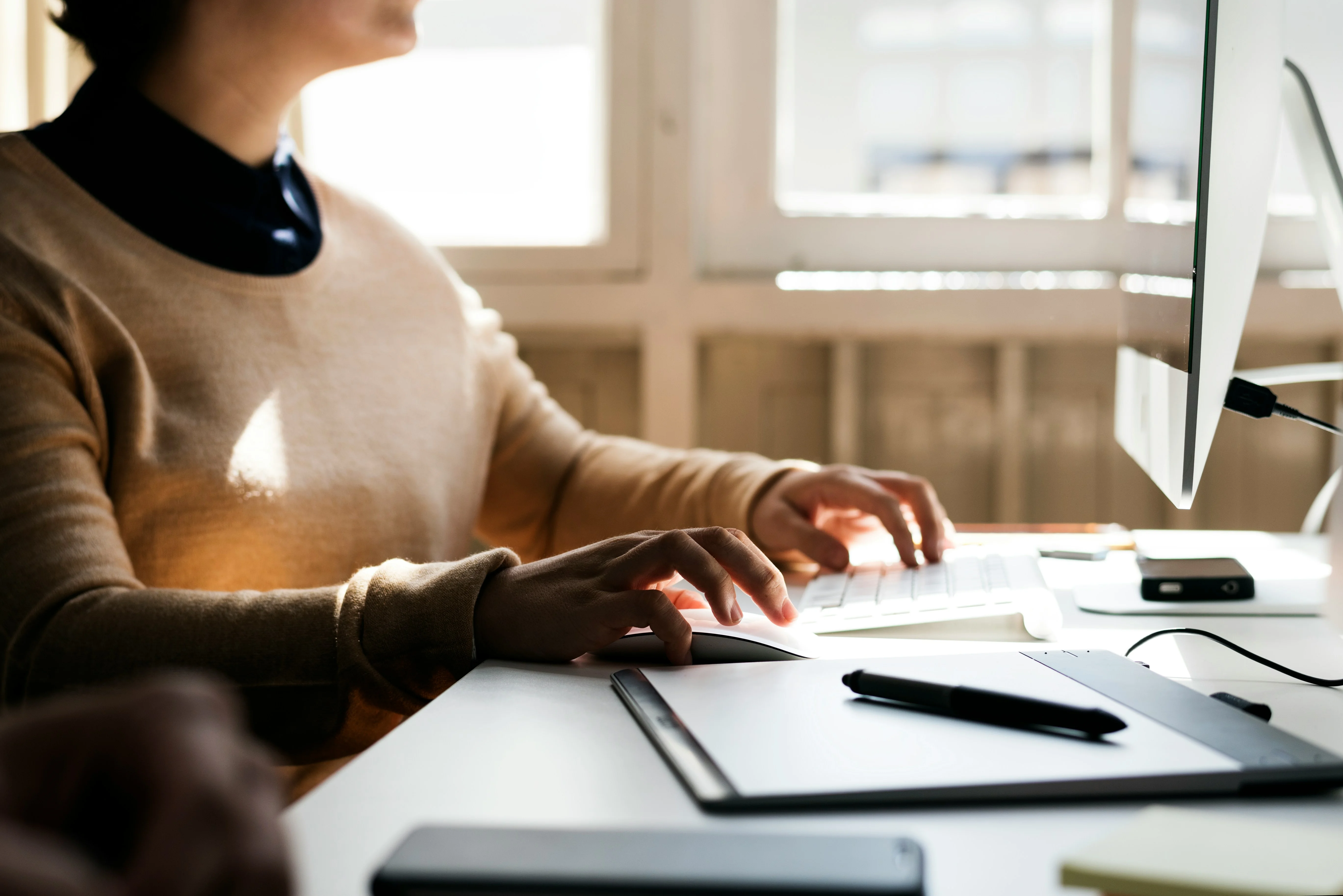A person in a beige sweater types on a keyboard at a desk, possibly analyzing market research with survey software. Sunlight streams through a window, illuminating a pen, notebook, and electronic devices. The individual’s face is partially out of frame.