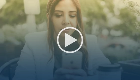 A woman with long hair is sitting outdoors at a café table, looking at her phone. A to-go coffee cup is on the table beside her. The image has a play button overlay, indicating its a video thumbnail.