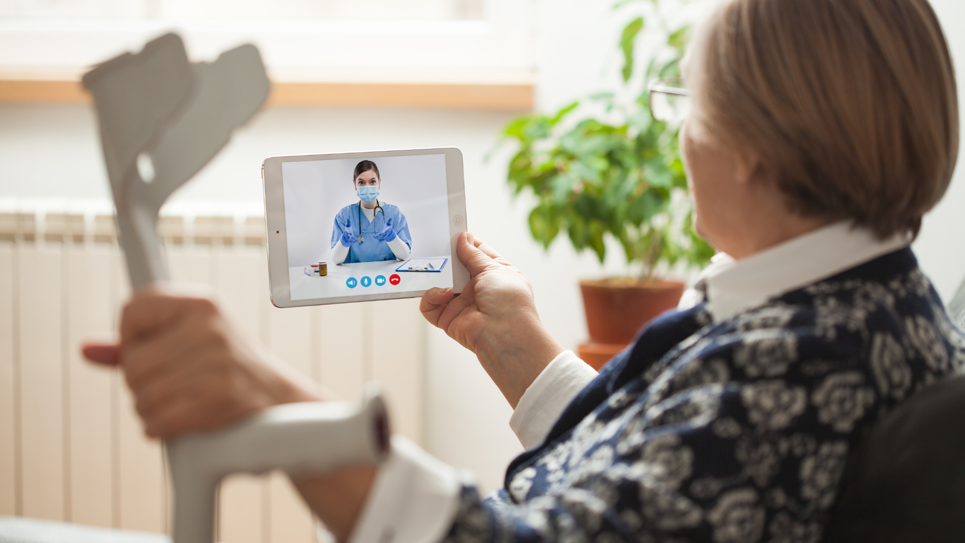 An older adult holding crutches watches a medical professional on a tablet during a video call. The person on the screen wears a blue uniform and a face mask. There is a potted plant in the background.