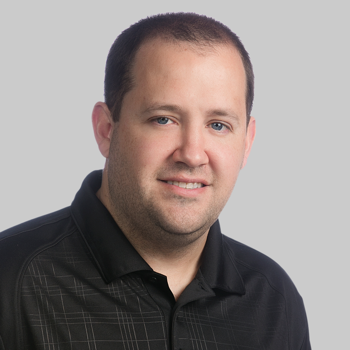 A man with short hair and a slight smile, Jason Pace from Alkami, is wearing a black collared shirt against a plain light gray background.