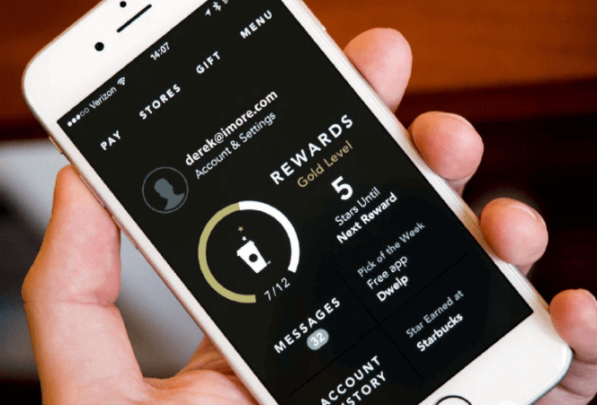 A hand holding a smartphone displaying Starbucks rewards app interface. The app shows a profile with a status of Gold Level and indicates 5 stars until the next reward. Theres an option to pick a free app of the week and a message count of 32.