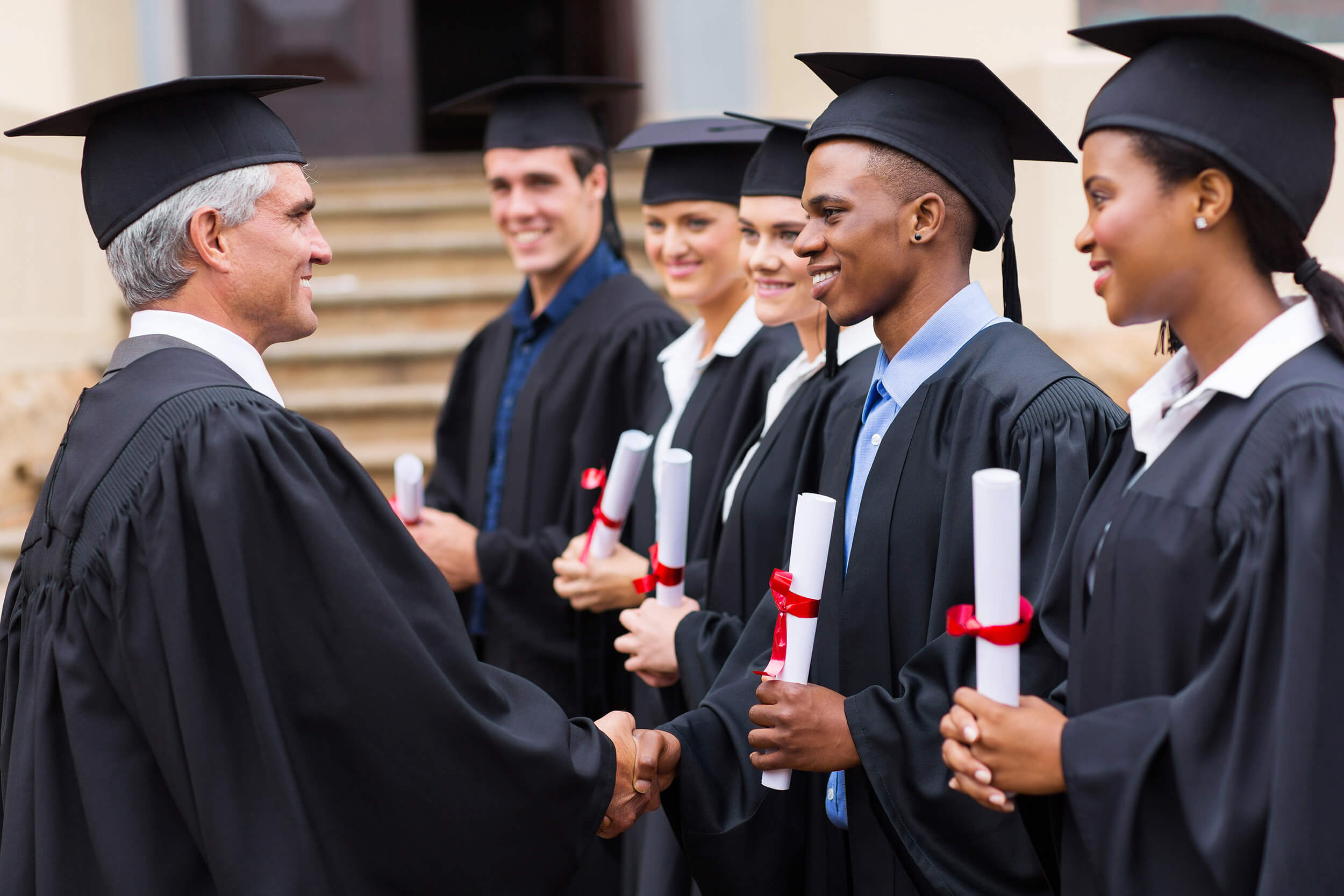 A line of graduates in caps and gowns receives diplomas from a faculty member. They are smiling and shaking hands in front of a building with steps. The graduates hold rolled-up certificates tied with red ribbons.