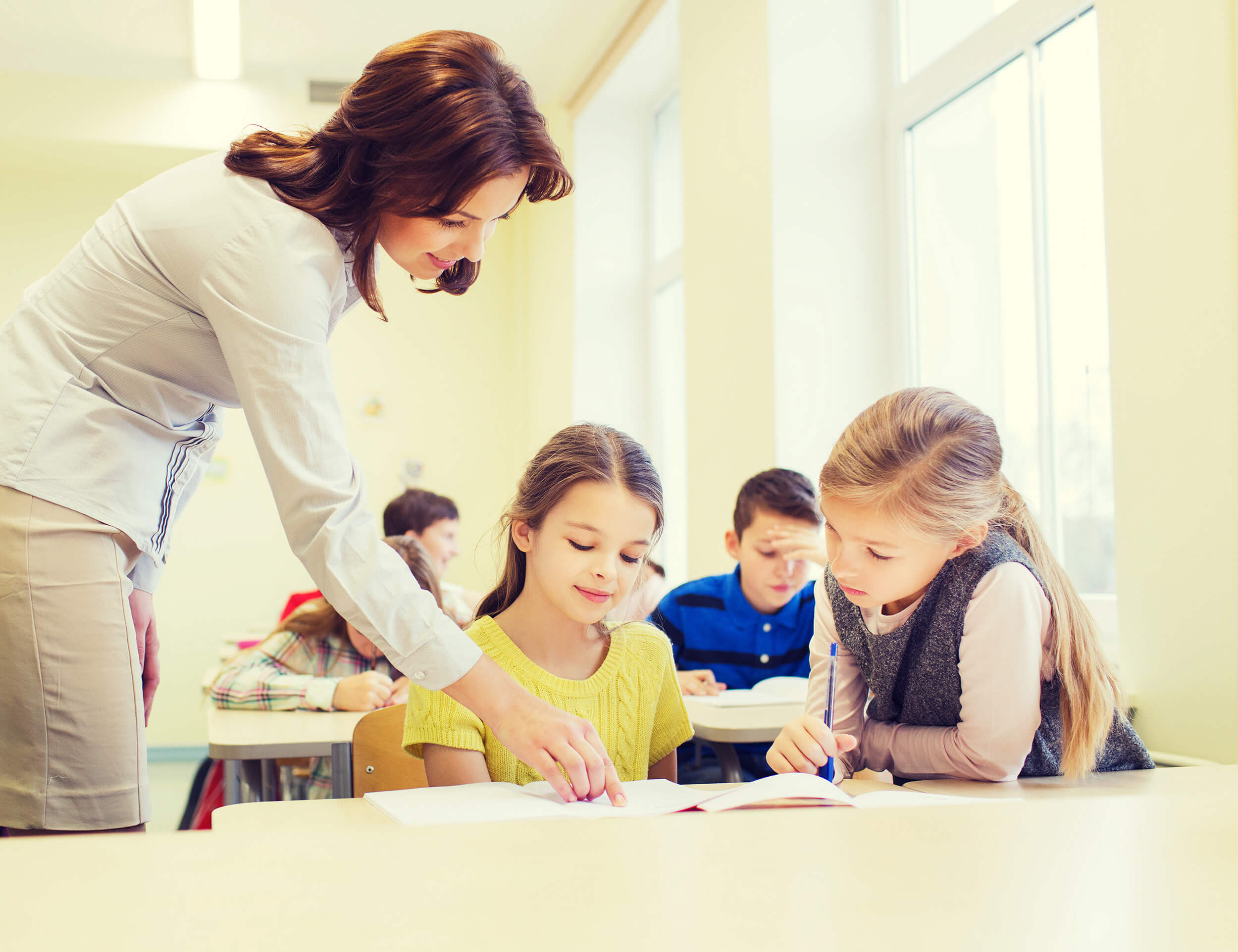 A dedicated teacher assists two young girls with their work in a bright classroom, while other students engage quietly at their desks near large windows.