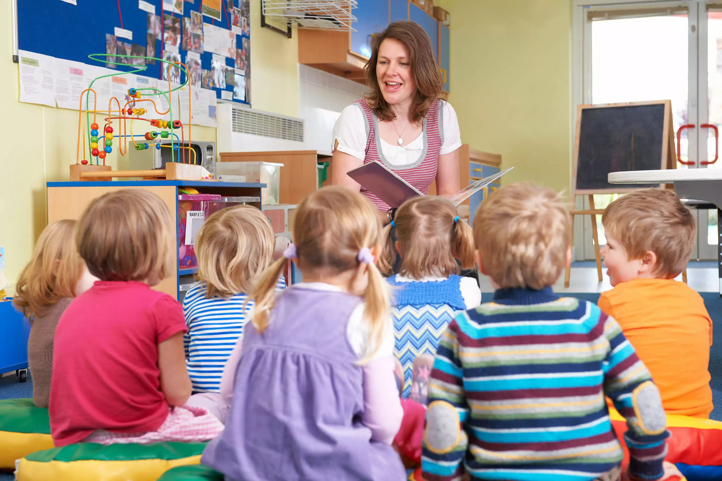 A teacher reads a book to a diverse group of young children sitting on colorful cushions in a classroom. The room is decorated with toys and educational materials. The children are attentively listening and facing the teacher.