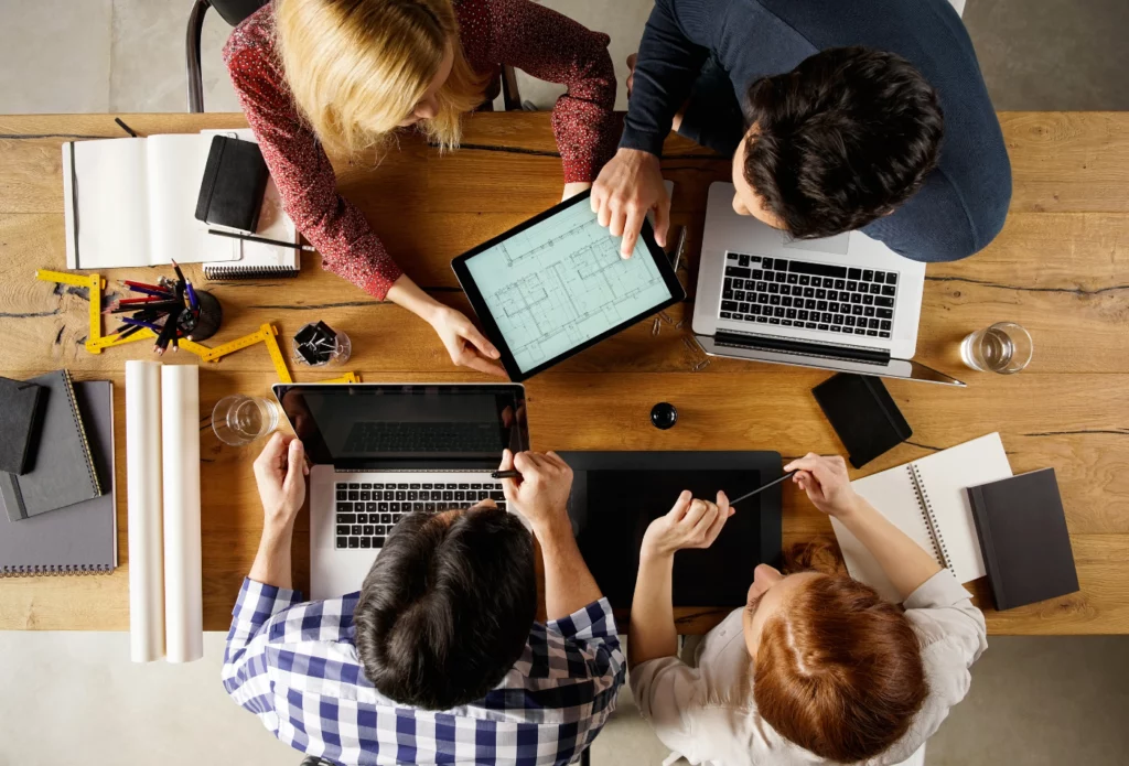 An overhead view of five people collaborating around a wooden table with laptops, tablets, notebooks, and papers. They appear to be discussing plans or designs on a digital tablet.