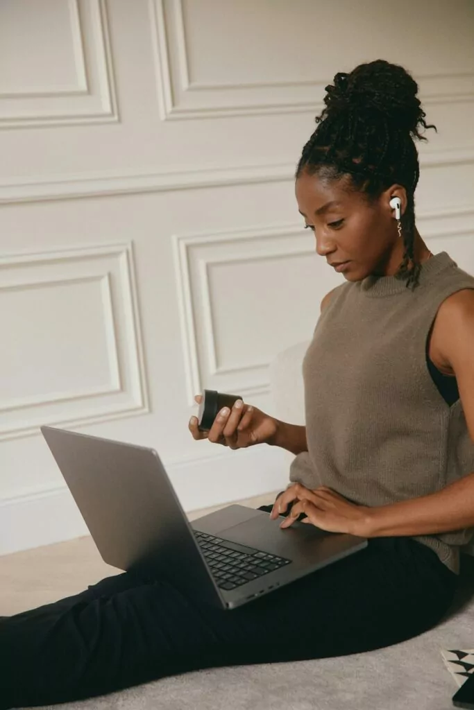 A woman with earbuds sits on a couch, focused on her laptop while holding a small black object, possibly a remote or charger. She is wearing a sleeveless top and black pants, with her hair pulled up in a bun. A white paneled wall is in the background.
