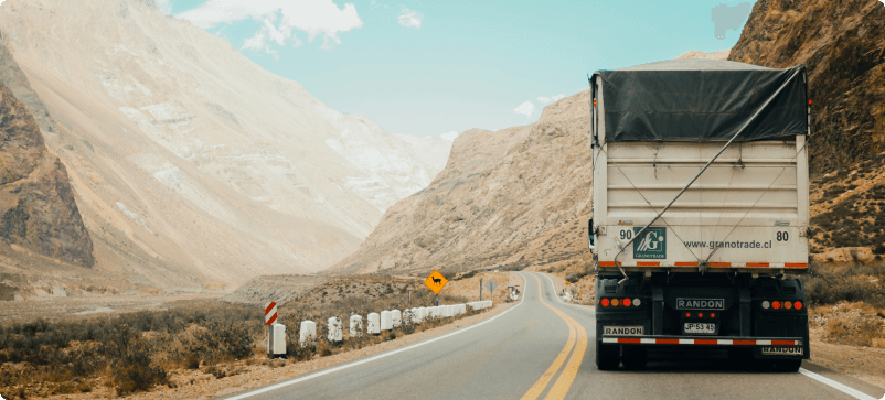A truck with a covered cargo bed drives along a winding mountain road. The landscape features rugged, rocky hills and sparse vegetation under a partly cloudy sky. Road signs and guard rails line the road.