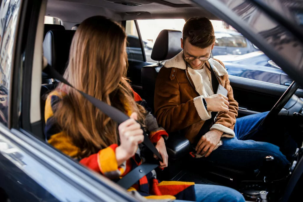 Young couple in a car fastening their seatbelts, demonstrating responsible driving habits. Man wearing brown jacket and woman in colorful sweater prioritizing safety before travel. Interior car shot highlighting the importance of seatbelt use and road safety awareness among young adults.