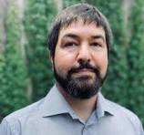 A man with short dark hair and a full beard, wearing a light gray collared shirt, stands in front of a blurred green background.
