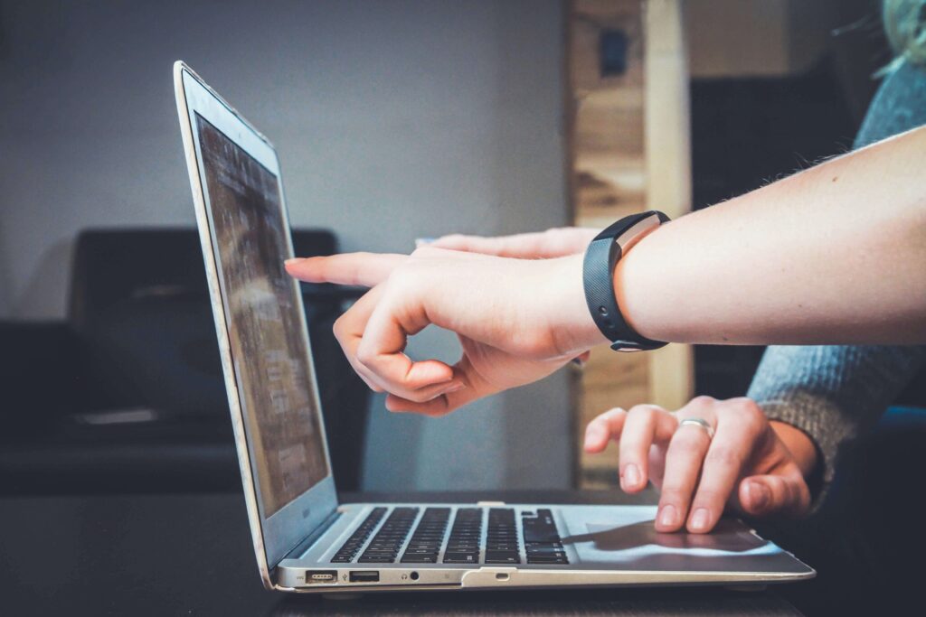 Two hands interacting with a laptop, one pointing at the screen and the other near the keyboard, suggesting collaboration to enhance retail customer experience. A blurred indoor setting provides context.