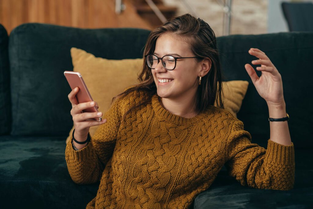 A person wearing glasses and a mustard-colored sweater is sitting on a dark green sofa, smiling while looking at their smartphone. A yellow pillow is in the background.