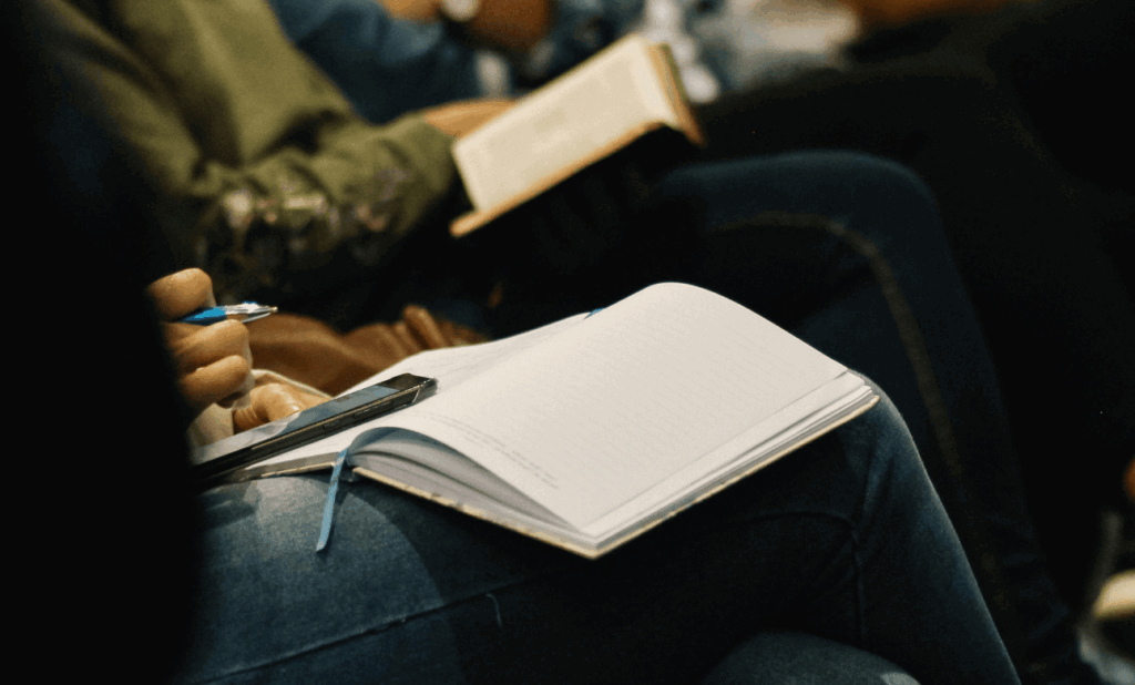 Photo of the torsos of people sitting in a row. Each person has a notebook in their lap, the person closest is scrolling on their phone with a pen in their hand. It looks like they are listening to a speaker