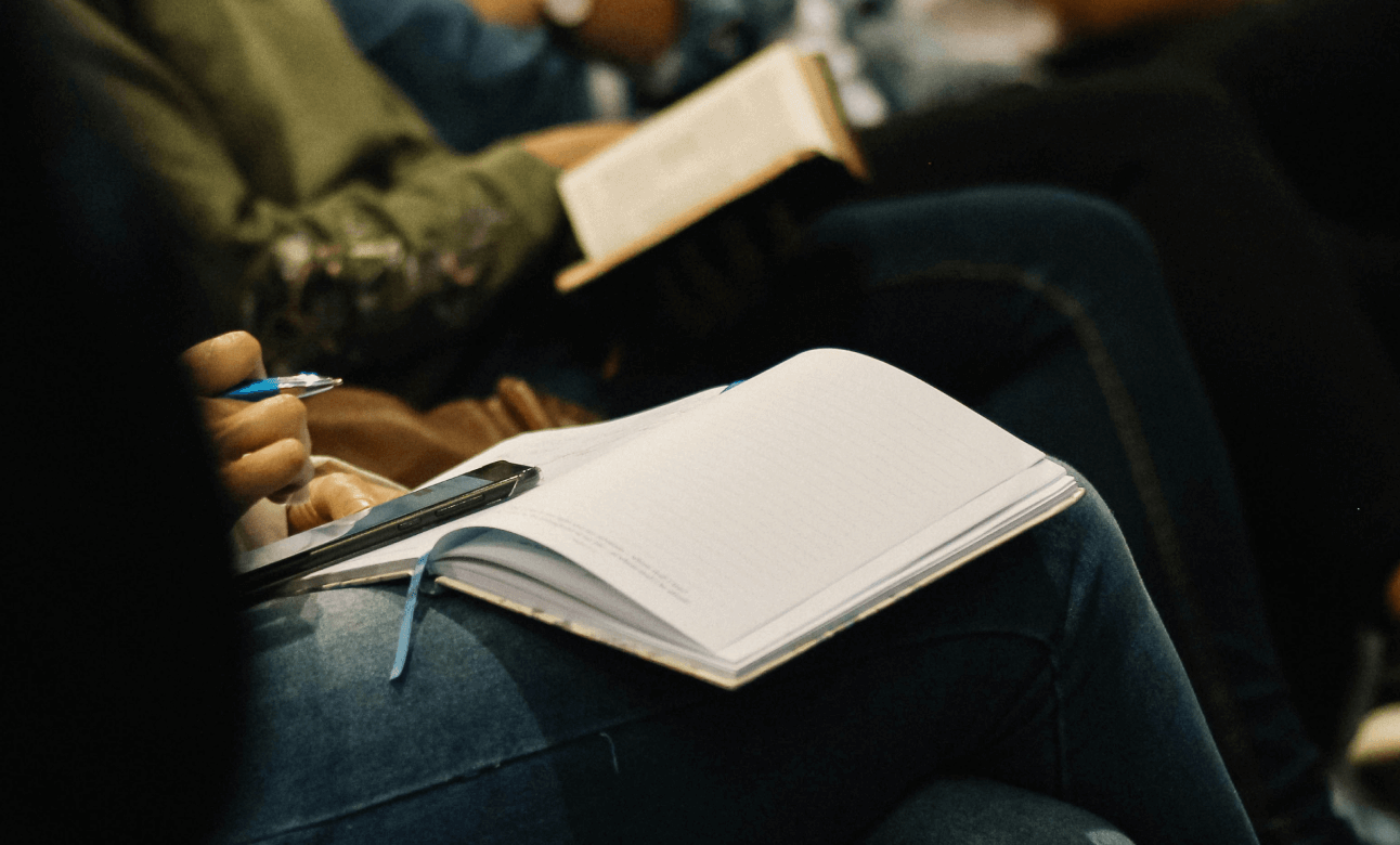 Photo of the torsos of people sitting in a row. Each person has a notebook in their lap, the person closest is scrolling on their phone with a pen in their hand. It looks like they are listening to a speaker