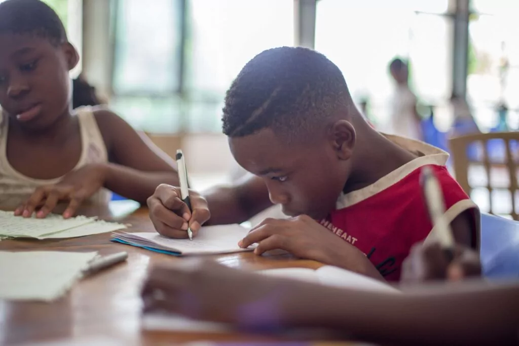 Three children are seated at a table, focused on writing in notebooks. The child in the red shirt is at the center, concentrating on his work. Soft, natural light fills the room through large windows in the background.