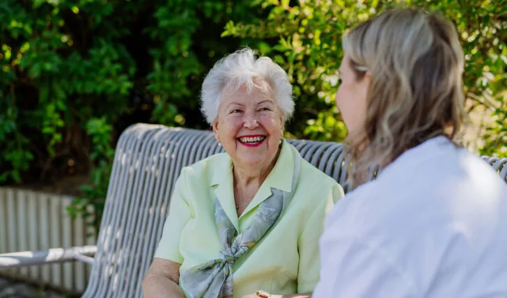 Elderly woman with white hair wearing light green blouse laughing while holding hands with caregiver on outdoor bench, surrounded by greenery