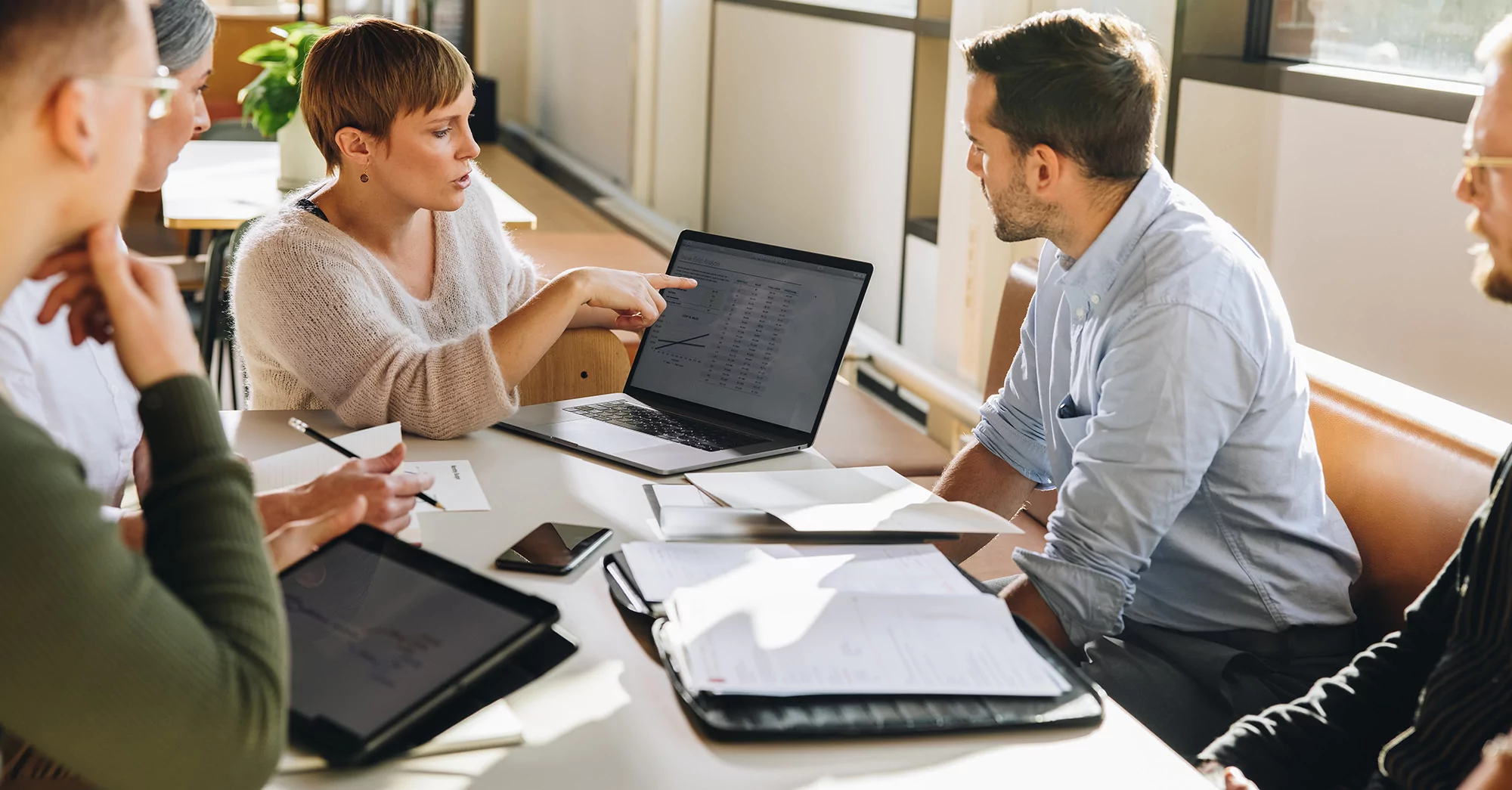 Four people sit around a table in a modern office, discussing ways to automate their CX processes. One person points at a laptop screen displaying graphs, with laptops and papers spread across the table.