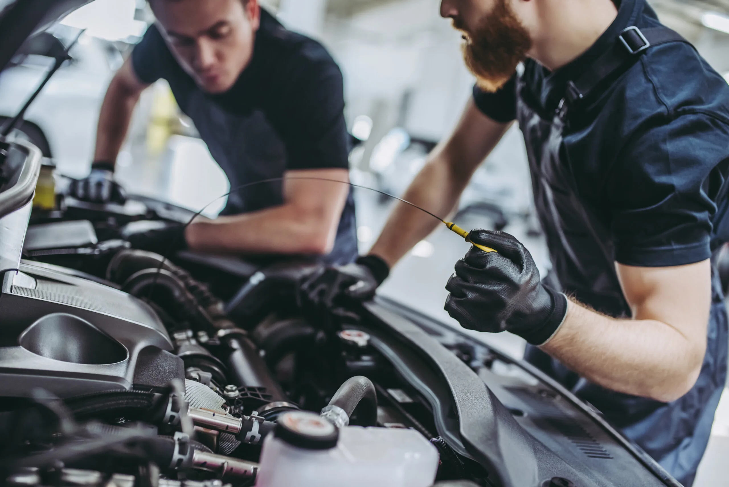 Two mechanics wearing gloves inspect a car engine; one holds a dipstick while the other leans over the open hood, focusing on the engine components.