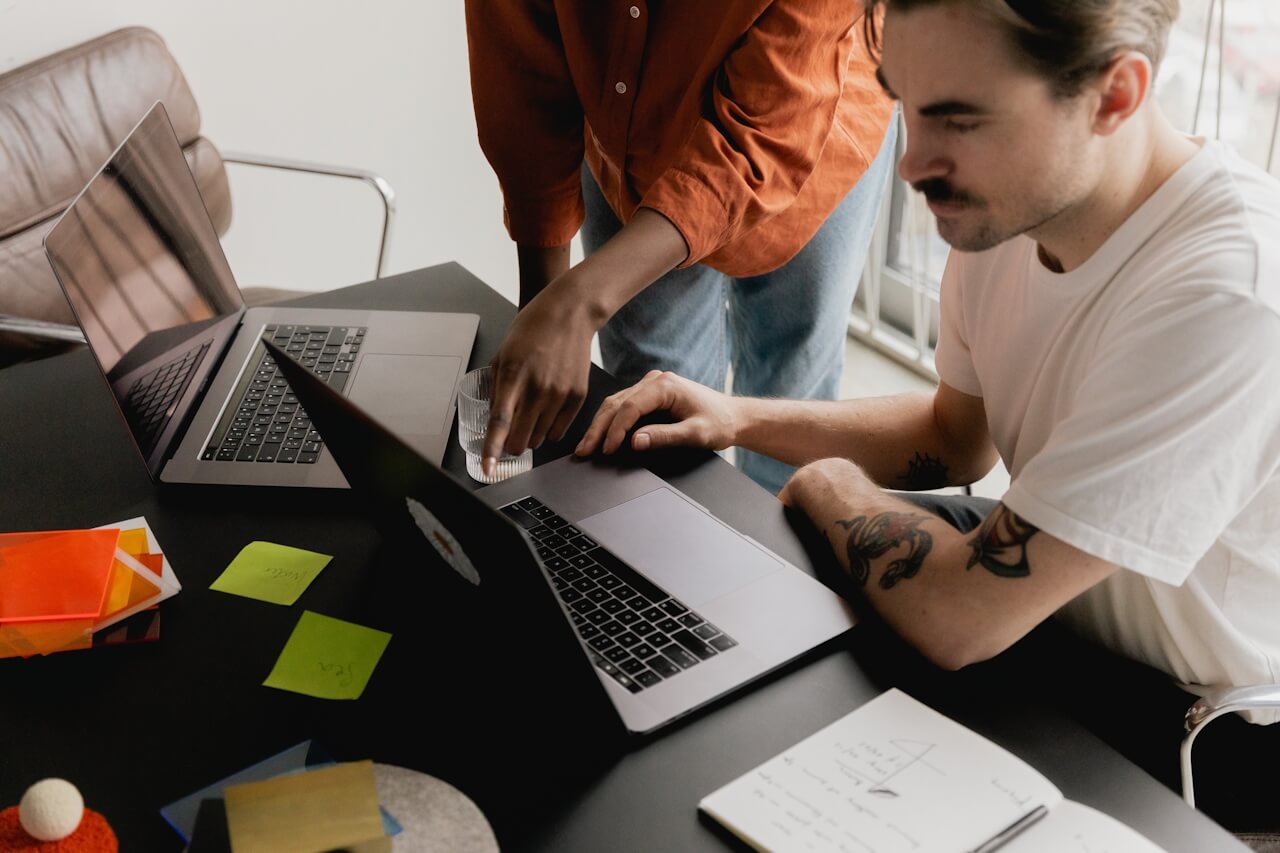 Two people working at a desk with laptops. One is seated, using a laptop and a notebook with sketches. The other is standing, pointing at the screen. Sticky notes and a glass of water are also on the desk.
