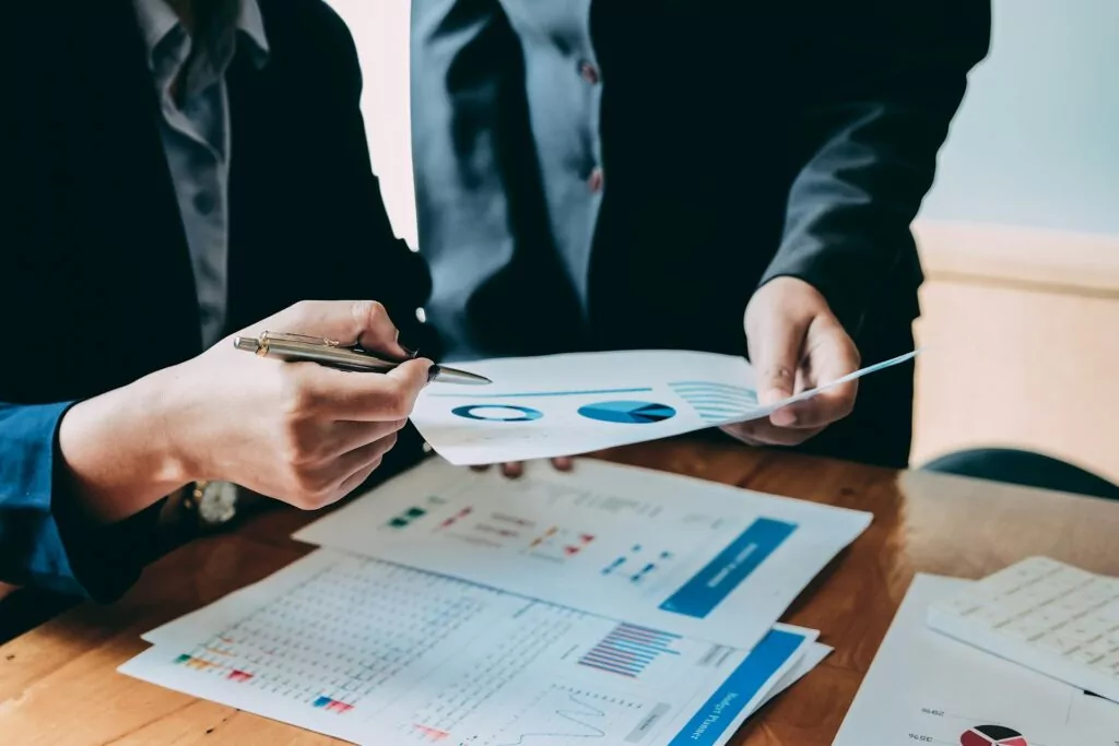 Two people in business attire review charts and graphs at a wooden desk, discussing data quality and analysis. One points to a document with a pen while the other holds a sheet with blue graphs, ensuring accuracy in their findings.