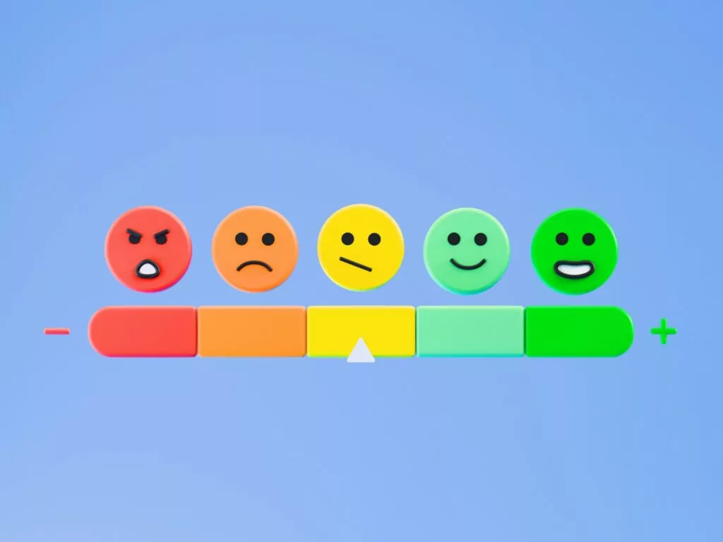 A horizontal scale used in NPS surveys with five colorful faces showing emotions from angry (red) to happy (green), a pointer on the neutral yellow face, and a minus sign on the left and plus sign on the right, set against a light blue background