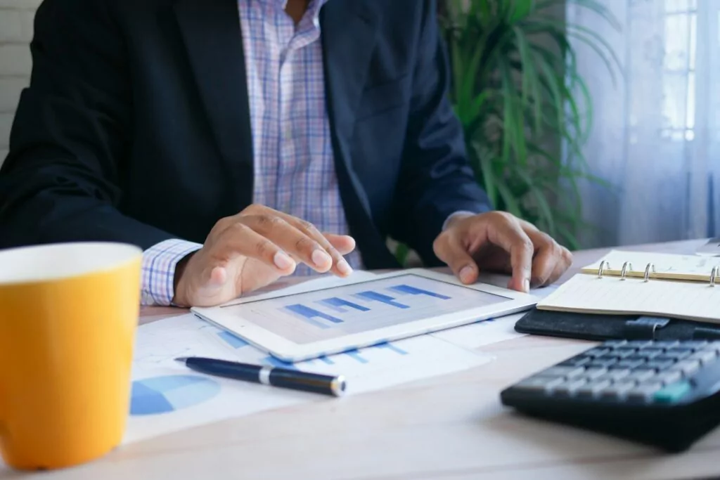 A person in business attire uses a tablet displaying bar charts at a desk with papers, a pen, a calculator, a notebook, and a yellow mug, with a plant in the background.