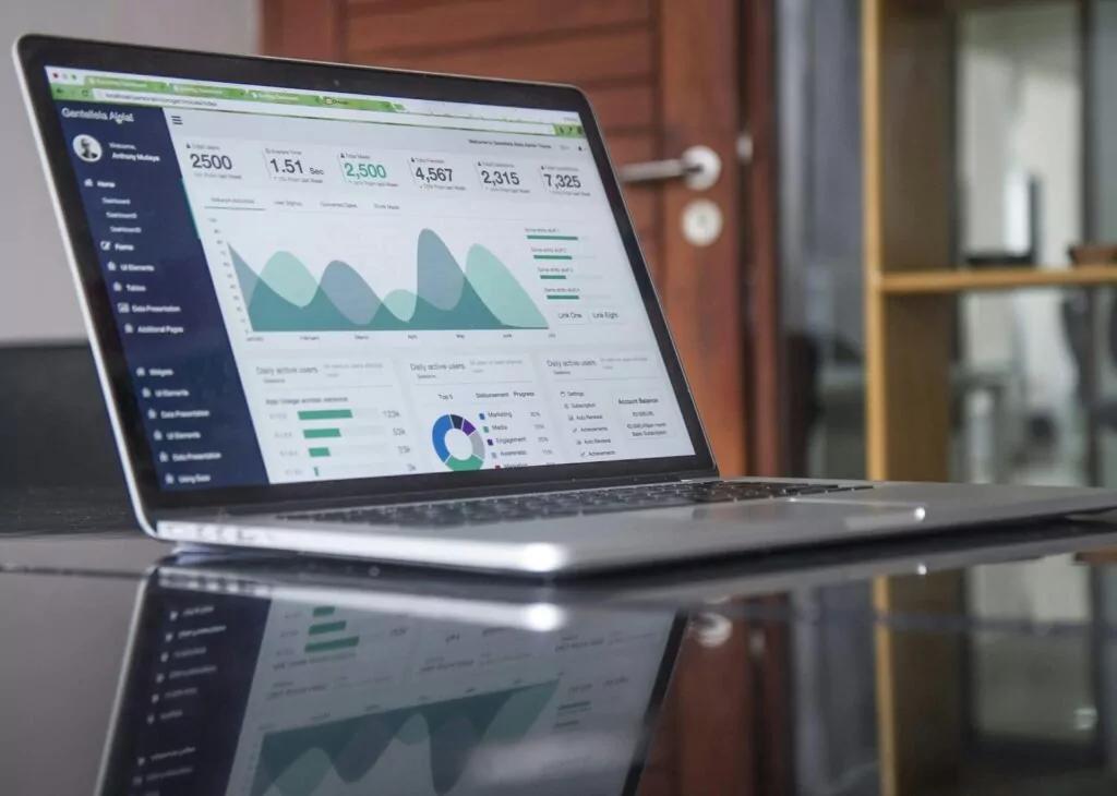 A laptop on a glass table displays a dashboard with graphs, charts, and cx measurement analytics in a modern office setting. The screen highlights various statistics and visual data representations.