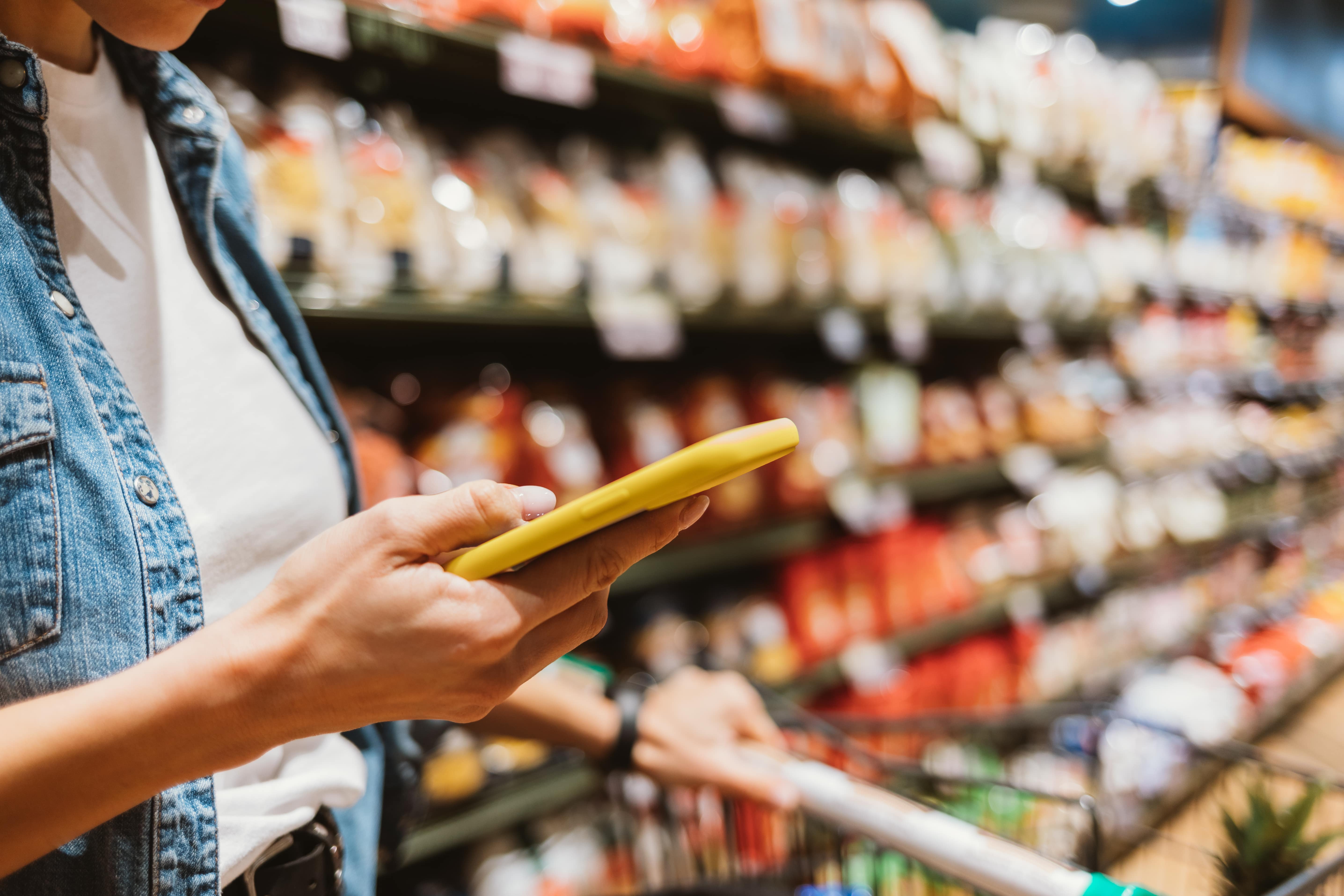 A person in a denim shirt uses a yellow smartphone while pushing a shopping cart through a grocery outlet aisle filled with various packaged food items.
