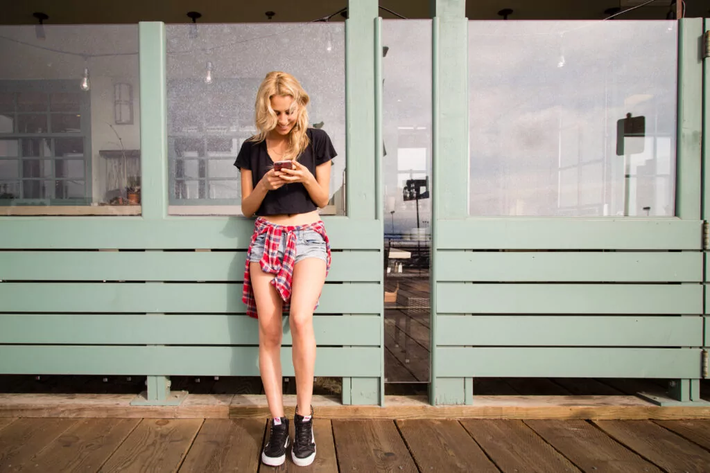 A young woman with blonde hair, wearing a black crop top, denim shorts, sneakers, and a plaid shirt tied around her waist, stands outside against a green wooden fence, reading a credit union case study on her phone.