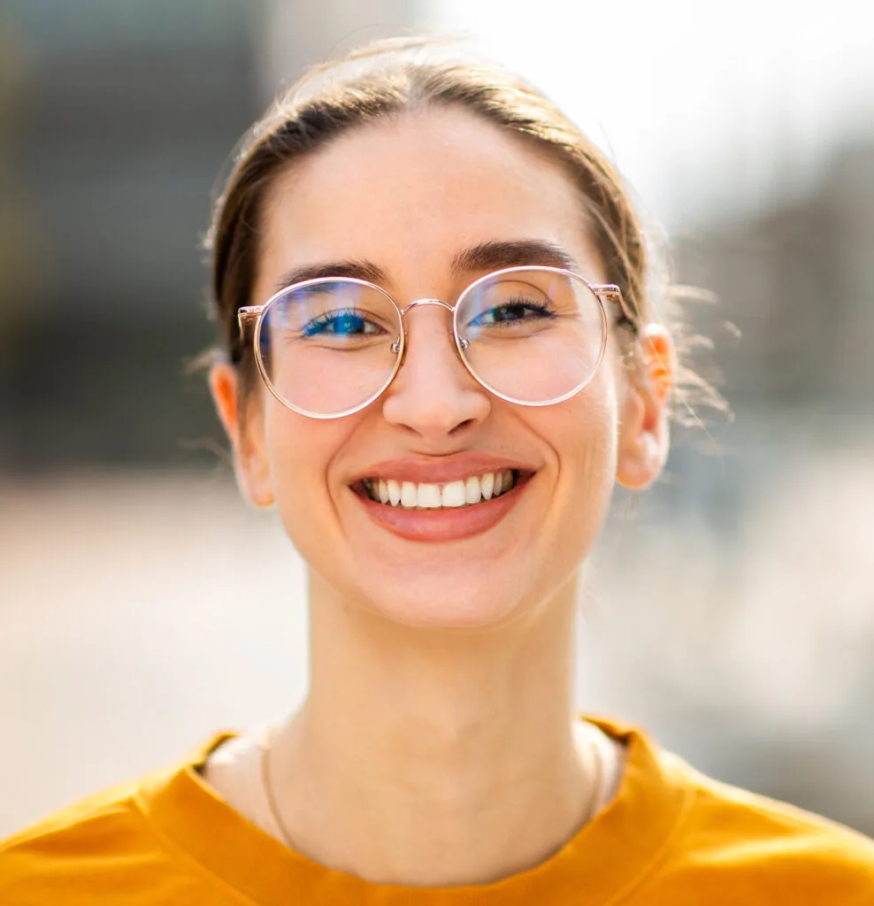 A young woman wearing round glasses and a mustard yellow top, smiles warmly at the camera. Her brown hair is pulled back and the background is softly blurred.