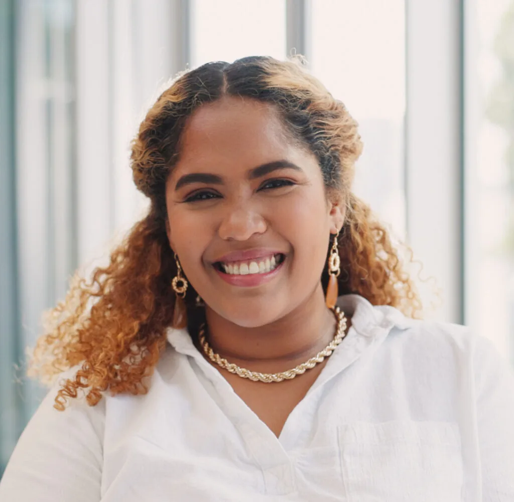 A smiling woman with curly hair wearing a white shirt, gold earrings, and a gold necklace, standing in front of large windows with natural light streaming in.
