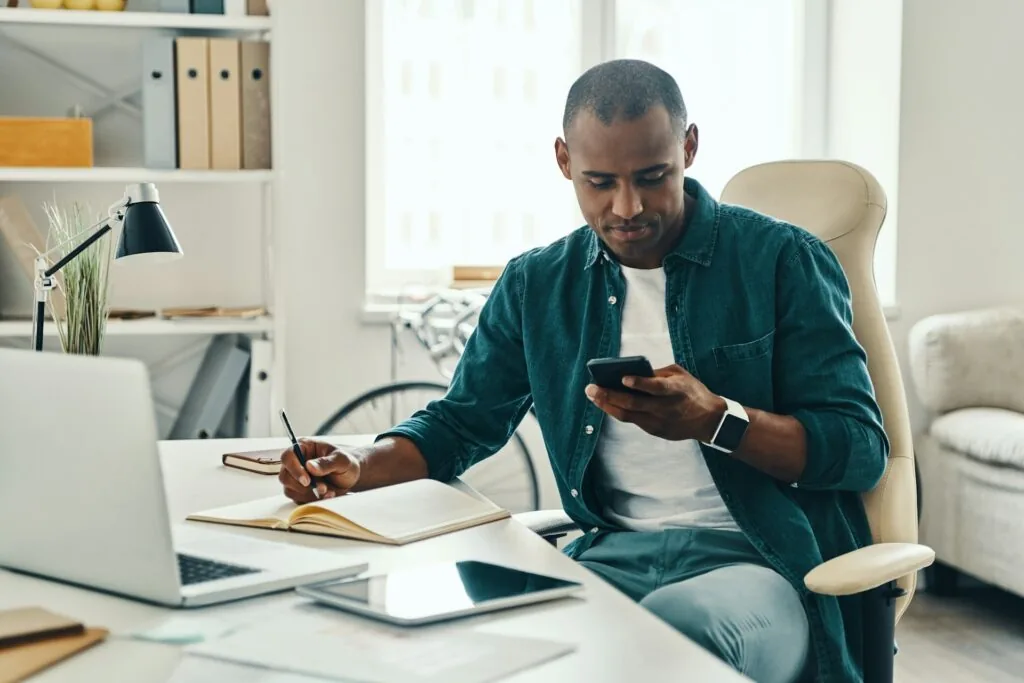 A man sits at a desk in a bright office, looking at his smartphone while holding a pen over an open notebook—seeking smarter digital feedback. A laptop, tablet, and documents are on the desk, with shelves and a window in the background.