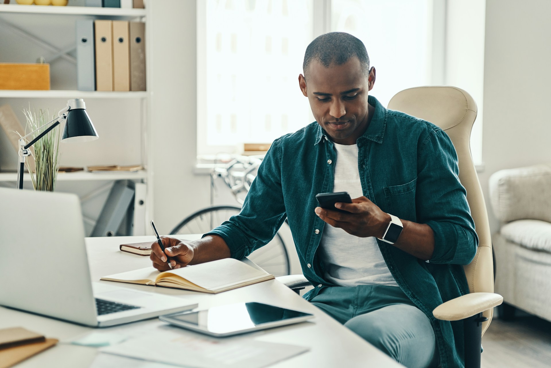 A man sits at a desk in a bright office, looking at his smartphone while holding a pen over an open notebook—seeking smarter digital feedback. A laptop, tablet, and documents are on the desk, with shelves and a window in the background.