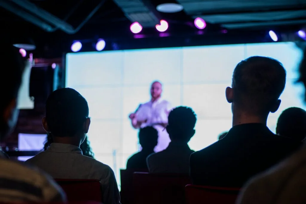 Audience members sit in a dark room facing a brightly lit stage, where an influencer is giving a presentation in front of a large digital screen. The presenter is blurred from the cameras perspective.