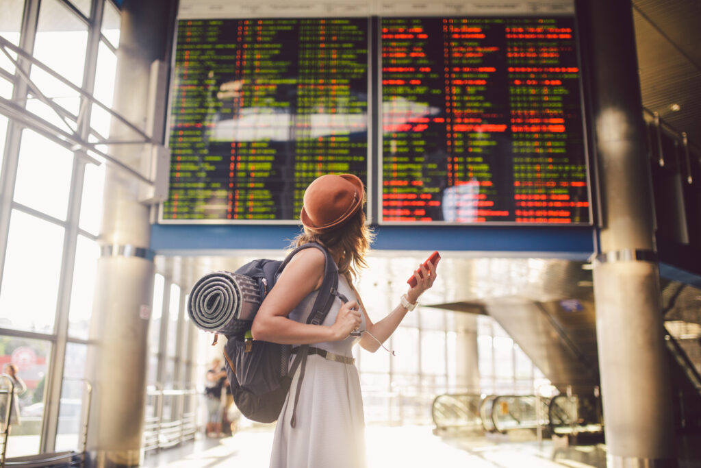 A woman wearing a hat and backpack looks up at a large digital departures board in an airport, holding her phone in one hand as sunlight streams through the windows, planning her journey on the c2c railway.