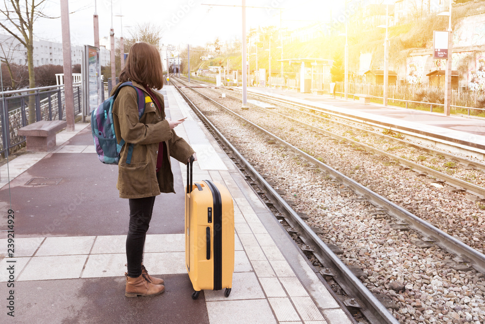 A person with a backpack stands on a c2c railway platform, holding a yellow suitcase and checking their phone. The tracks are empty, and the sun is shining brightly in the background.