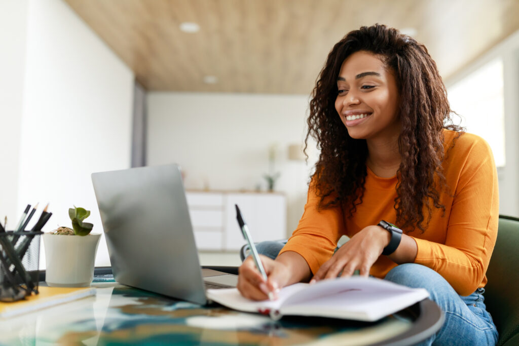 A woman with curly hair, wearing an orange top, sits at a table, smiling while looking at a laptop and writing in a notebook with a pen. The room appears bright and modern.