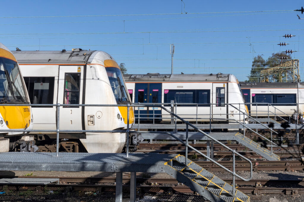 Several modern passenger trains are parked on parallel tracks at a train yard, with metal walkways and stairs providing access. Overhead electric wires and a clear blue sky are visible in the background.
