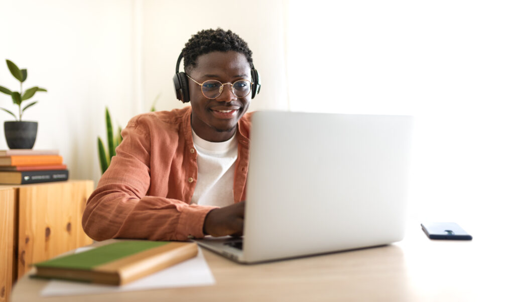 A man wearing headphones and glasses smiles while using a laptop at a desk. There are books, papers, a plant, and a smartphone nearby in a bright, cozy room.