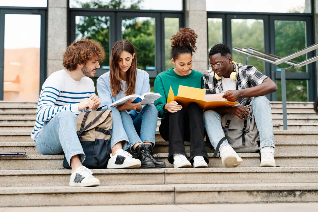 Four students sit on outdoor steps, reading books and papers together. They appear focused and are casually dressed, with backpacks nearby, suggesting a study session or group project.