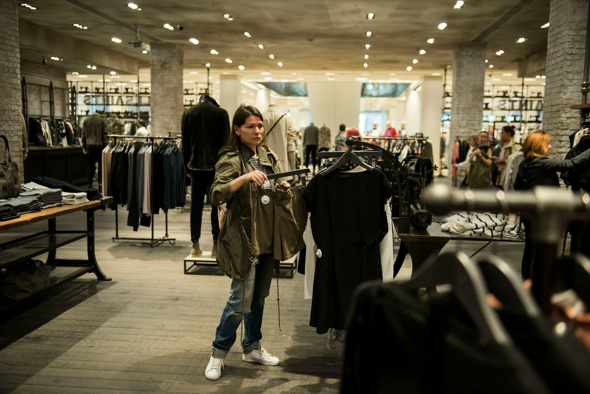 A woman stands in a clothing store, holding up a hanger with a dress. She appears to be shopping, surrounded by racks of clothes in a spacious, well-lit store with a modern industrial design.