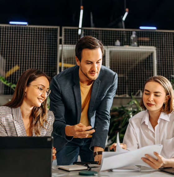 Three people sit at a table collaborating on paperwork. One man stands between two women, pointing at documents they are reviewing together. A laptop and notebook are on the table in a modern office setting.