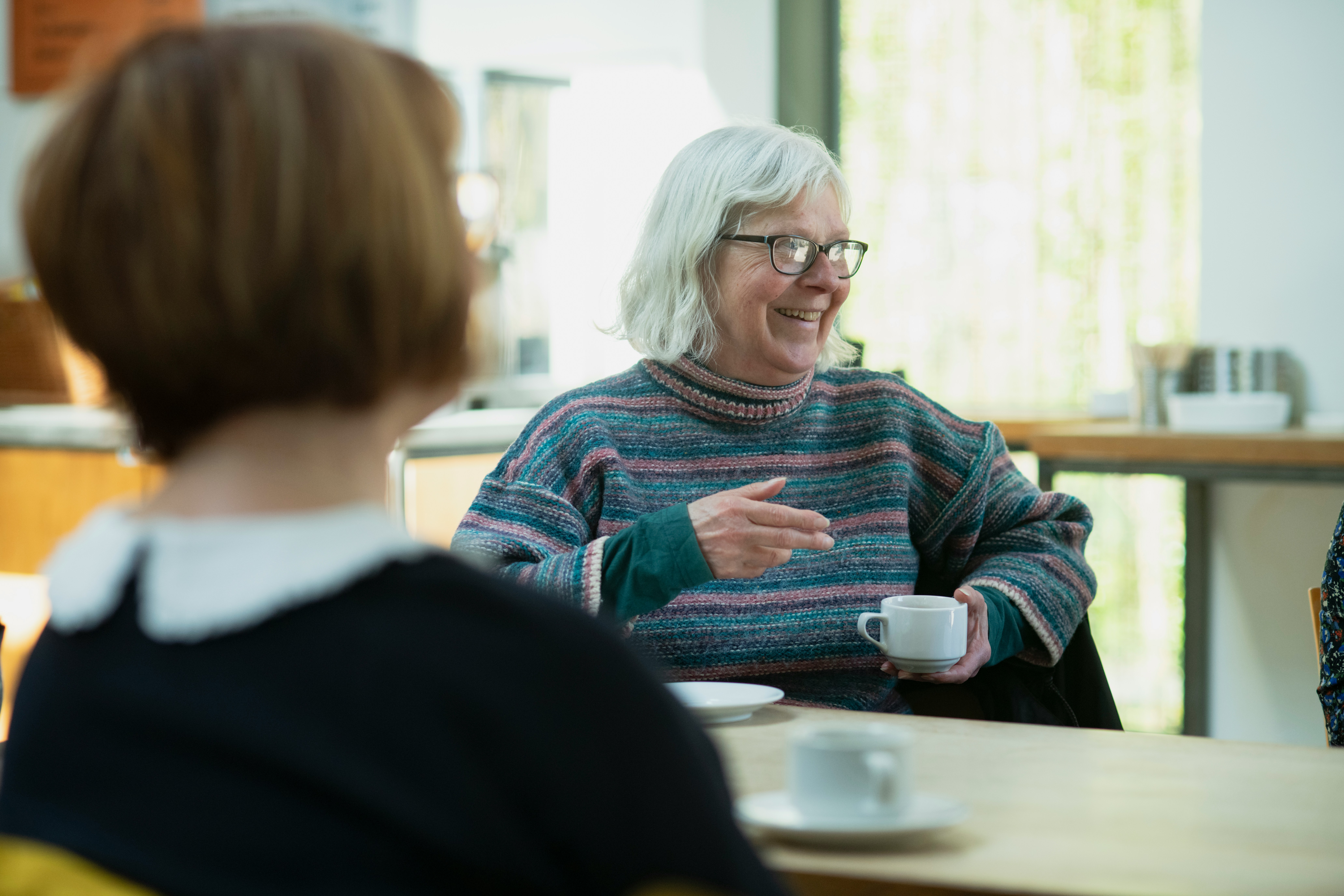An older woman with glasses and gray hair, wearing a striped sweater, smiles and gestures while holding a coffee cup at a table in a bright café. Another person is seated in the foreground.