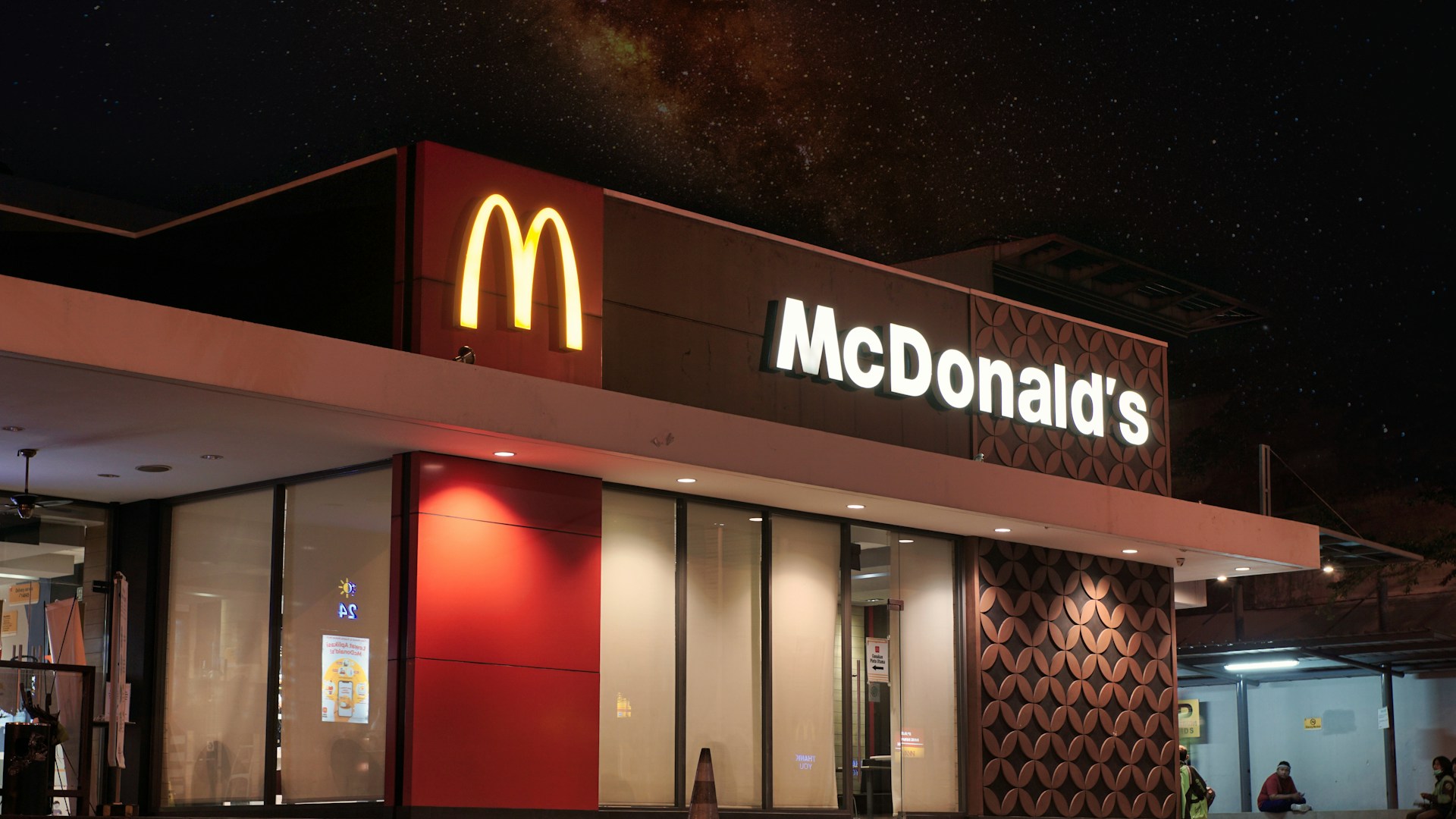 A McDonald’s restaurant at night, with the yellow “M” logo and illuminated “McDonald’s” sign on the building. The sky above shows stars and a hint of the Milky Way.
