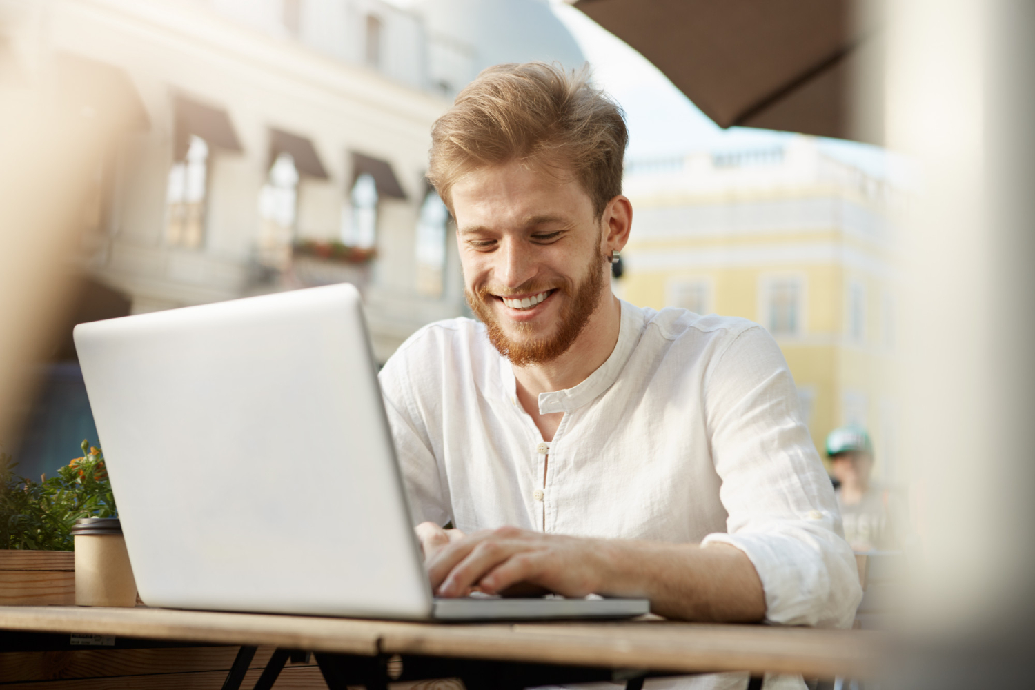 A smiling man with a beard, wearing a white shirt, sits at an outdoor café table using a laptop. The background shows blurred buildings and sunlight.