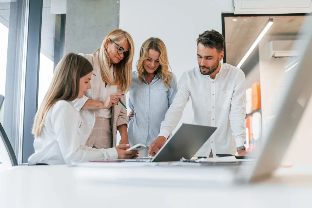 Four people in business attire gather around a desk, discussing work. One woman sits with a laptop, while the others stand, looking at the screen and documents, collaborating in a bright, modern office.
