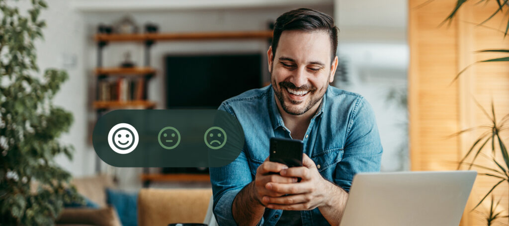 Smiling man using a smartphone at home beside a laptop, with a customer satisfaction rating graphic showing positive, neutral, and negative feedback options.