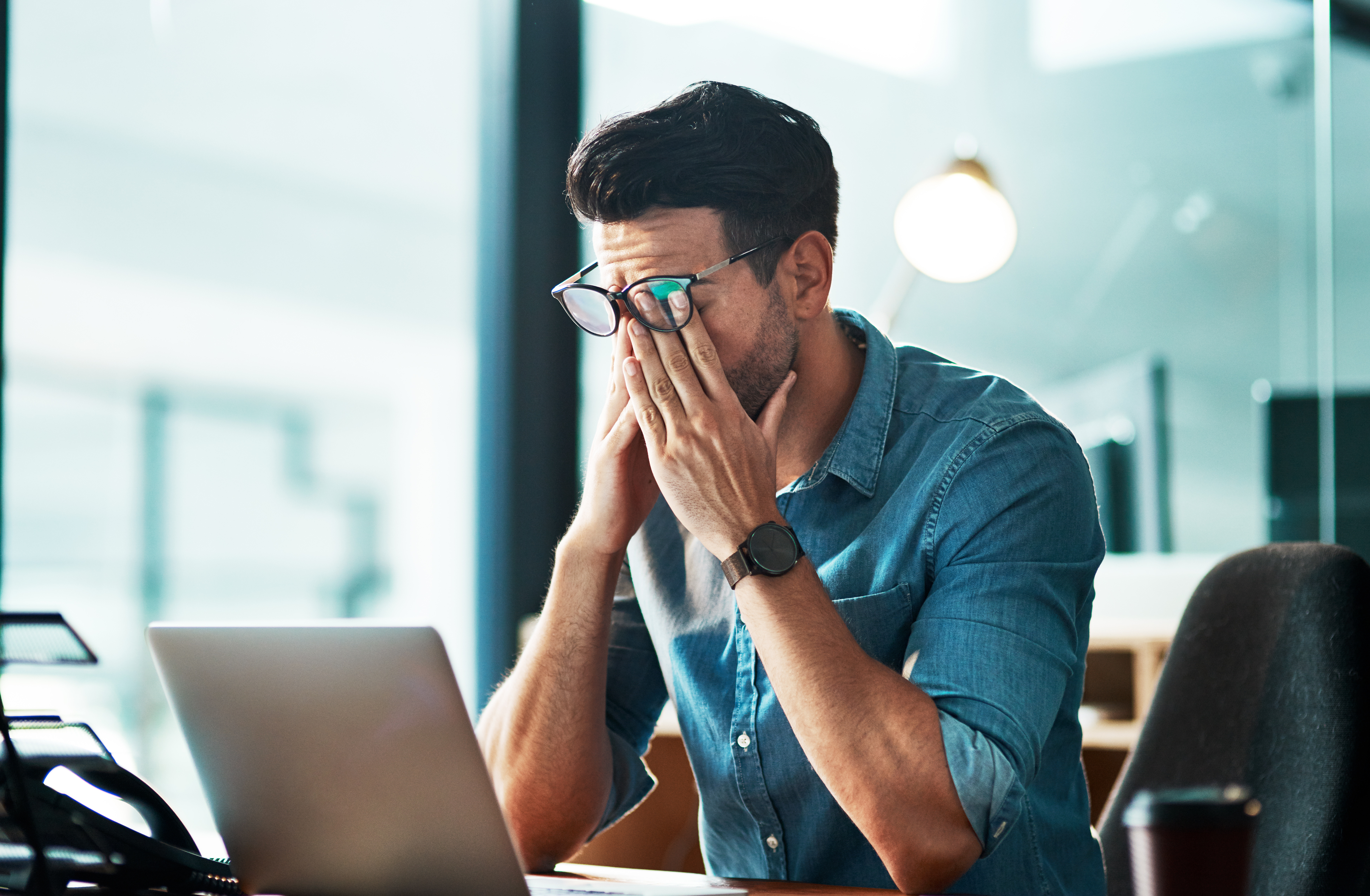 A man wearing glasses and a denim shirt sits at a desk with a laptop, rubbing his eyes in apparent fatigue or stress. The background shows an office setting with a blurred lamp and window.