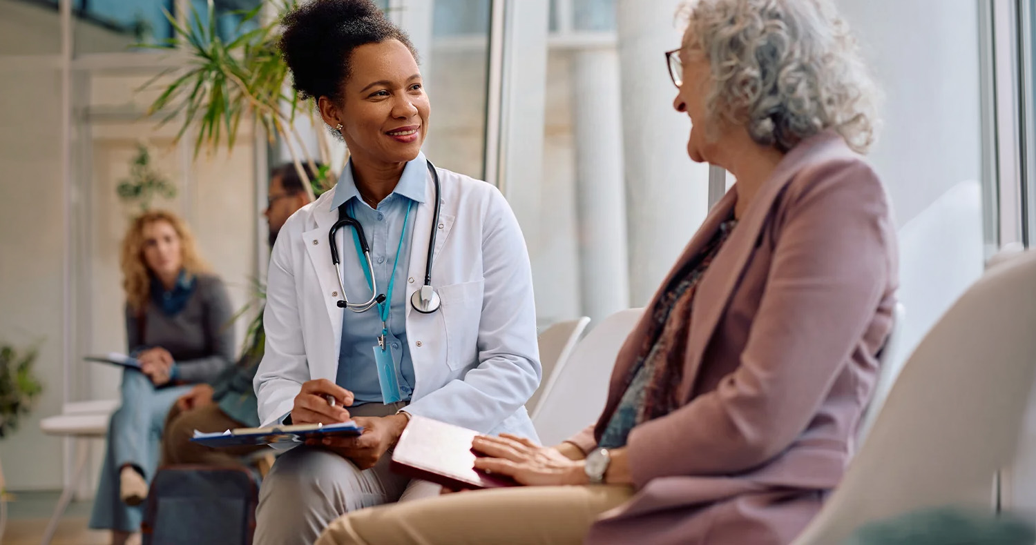 A doctor with a stethoscope smiles and talks with an older woman in a waiting area, both seated and holding clipboards, with other people visible in the background.